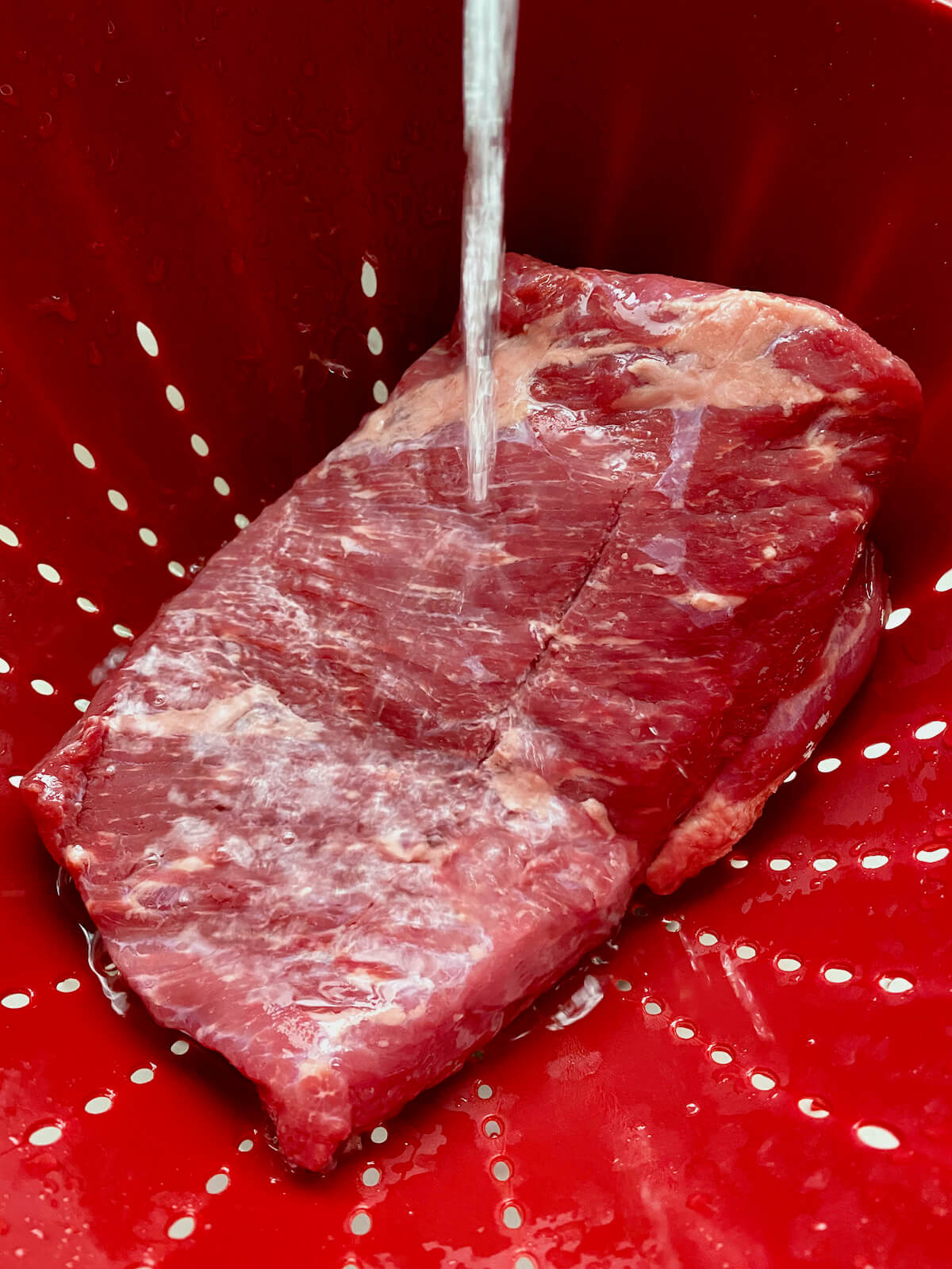 Corned beef being rinsed under running water in a red colander.