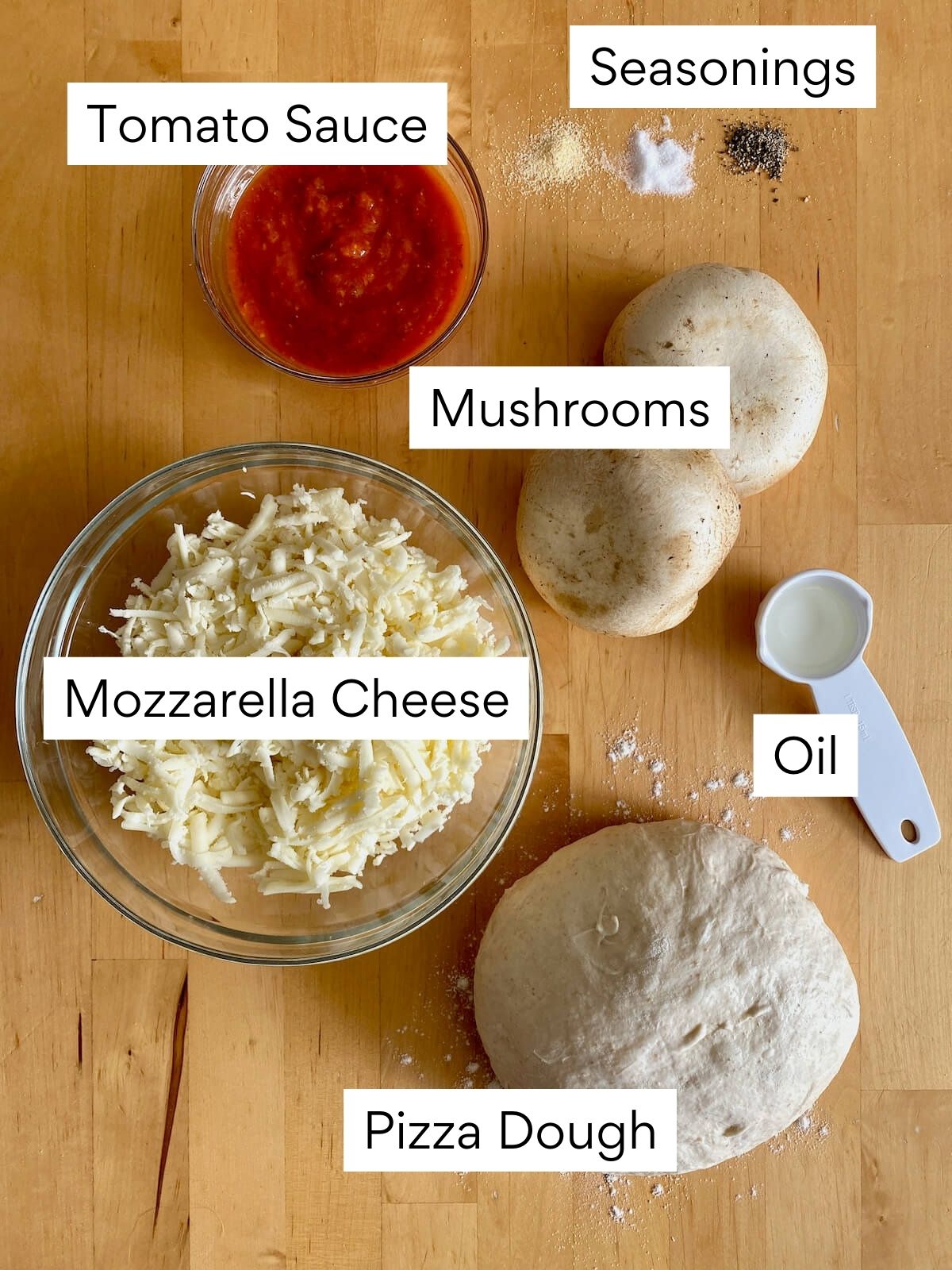 The ingredients to make mushroom pizza on a butcher block countertop. Each ingredient is labeled with text. They include tomato sauce, seasonings, mushrooms, mozzarella cheese, oil, and pizza dough.