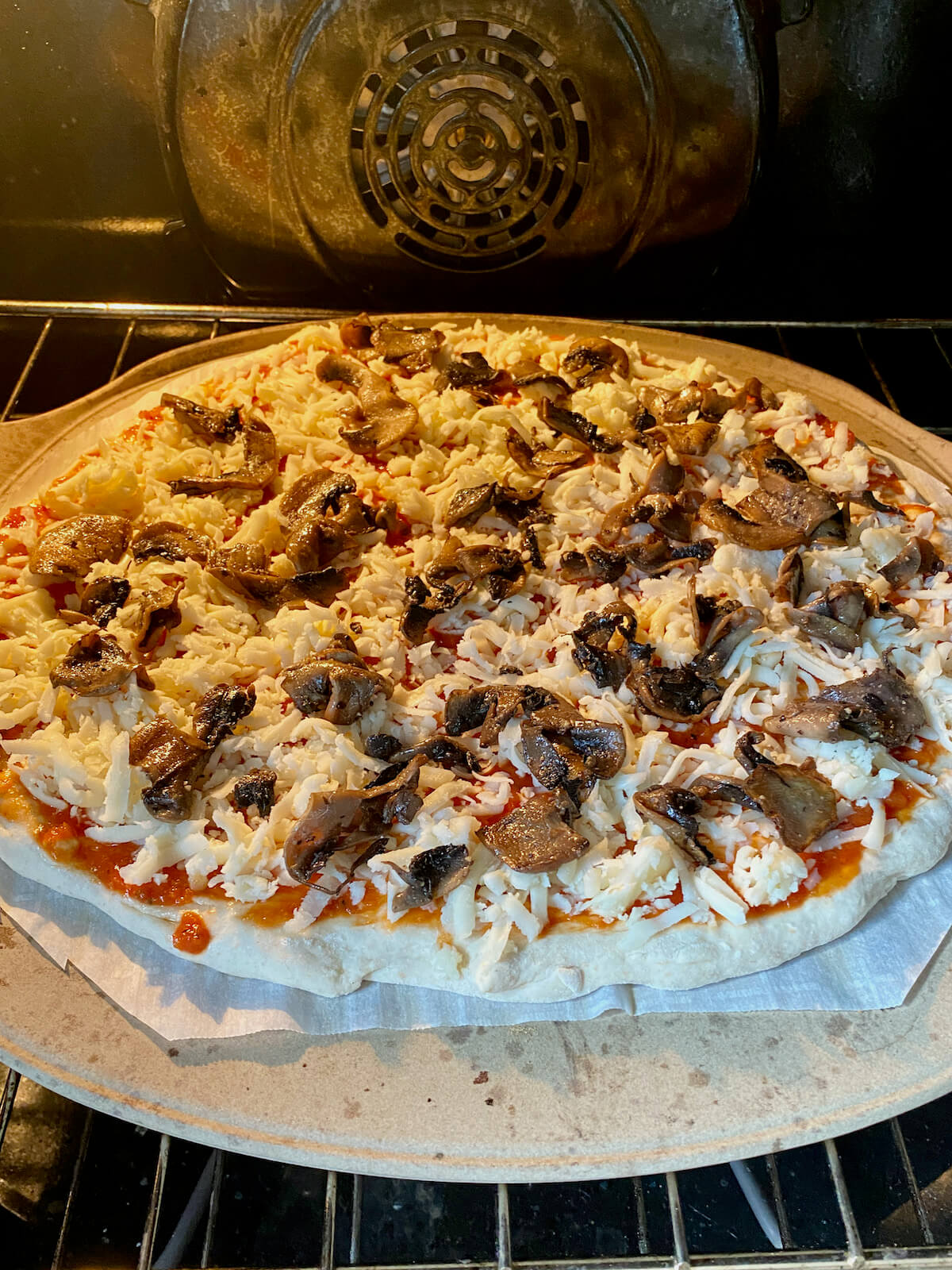Mushroom pizza on a piece of parchment paper on a pizza stone inside of a hot oven.