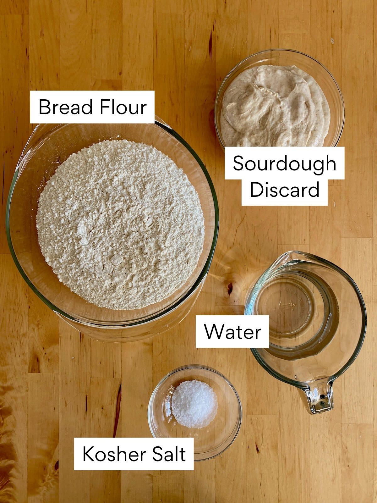 The ingredients to make sourdough discard pizza dough on a butcher block countertop. Each ingredient is labeled with text. They include bread flour, sourdough discard, water, and kosher salt.