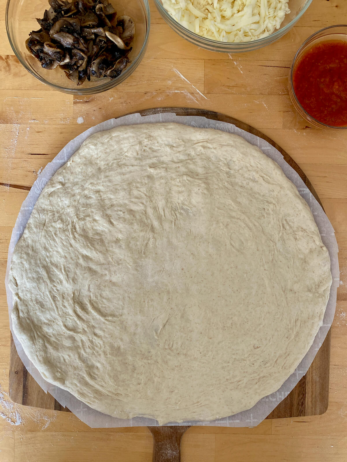 Sourdough discard pizza dough stretched out on a parchment-lined pizza peel. The stretched dough is surrounded by bowls of ingredients such as cooked mushrooms, shredded mozzarella cheese, and tomato sauce.