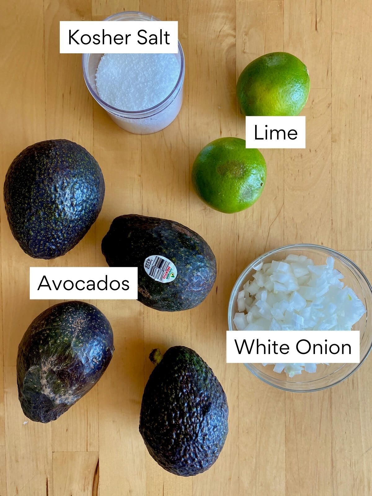 The ingredients to make 4-ingredient guacamole. Each ingredient is labeled with text. They include avocados, white onion, lime, and kosher salt.