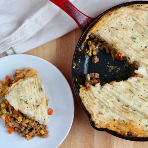 A cast iron skillet filled with ground turkey shepherd's pie. A single slice of the dish is missing from the skillet and is on a small white plate to the left.