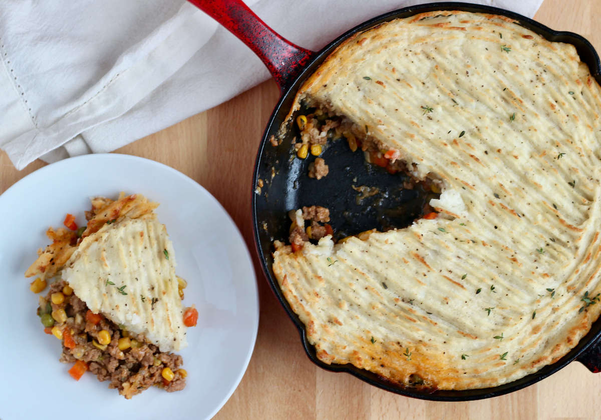 A cast iron skillet filled with ground turkey shepherd's pie. A single slice of the dish is missing from the skillet and is on a small white plate to the left.