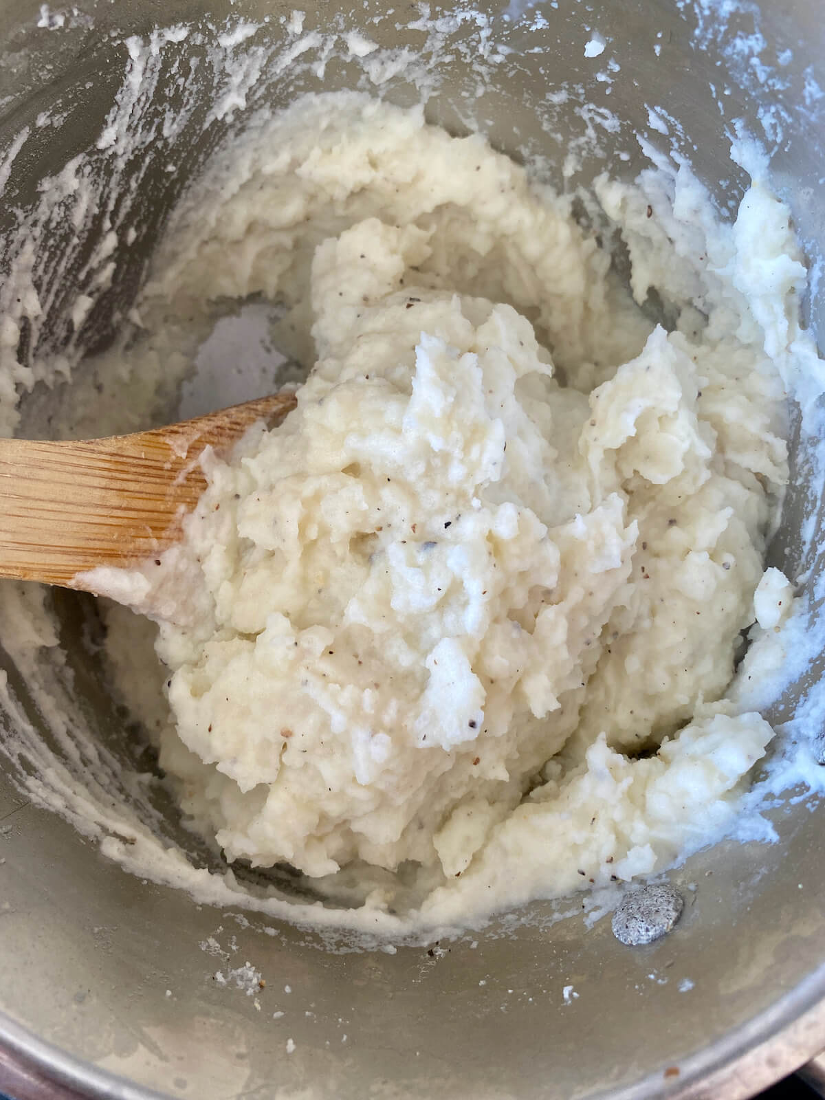 Mashed potatoes in a stainless steel pot being mixed by a wooden spoon.