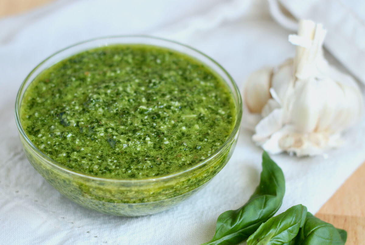 A bowl of basil pesto without pine nuts next to a head of garlic and a few basil leaves.