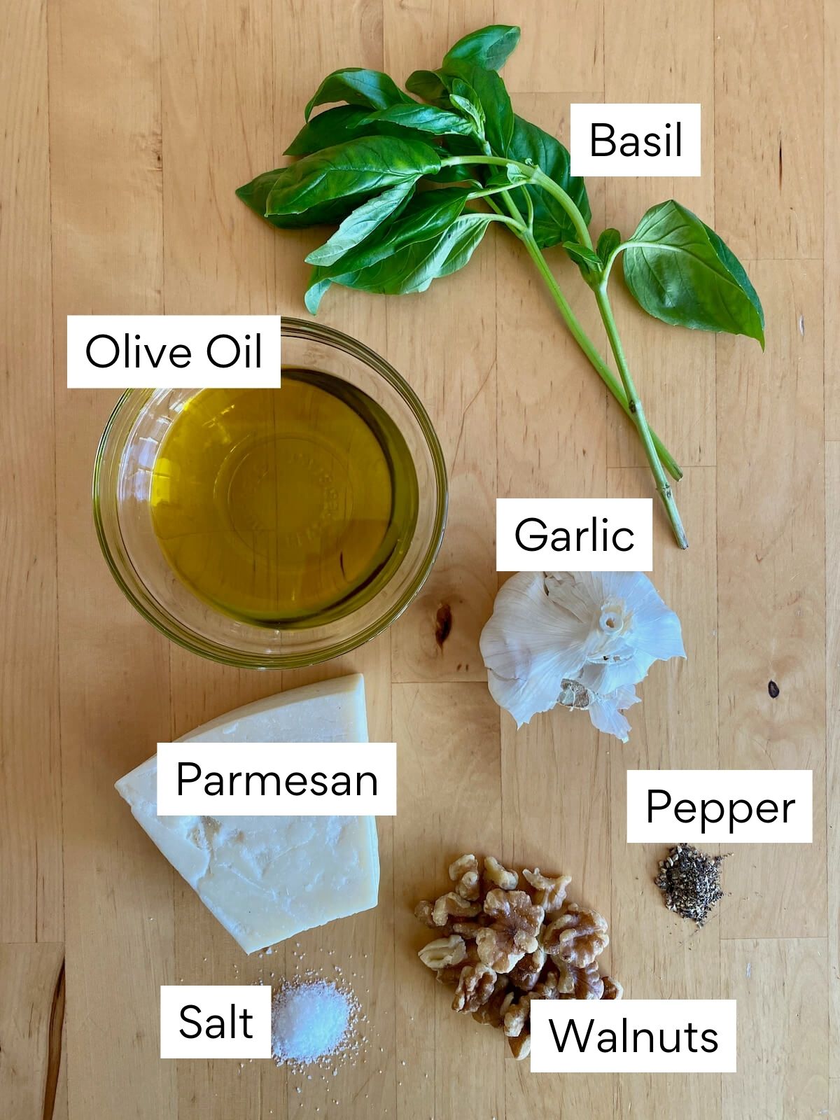 The ingredients to make pesto without pine nuts. Each ingredient is labeled with text. They include basil, olive oil, garlic, parmesan, walnuts, salt, and pepper.