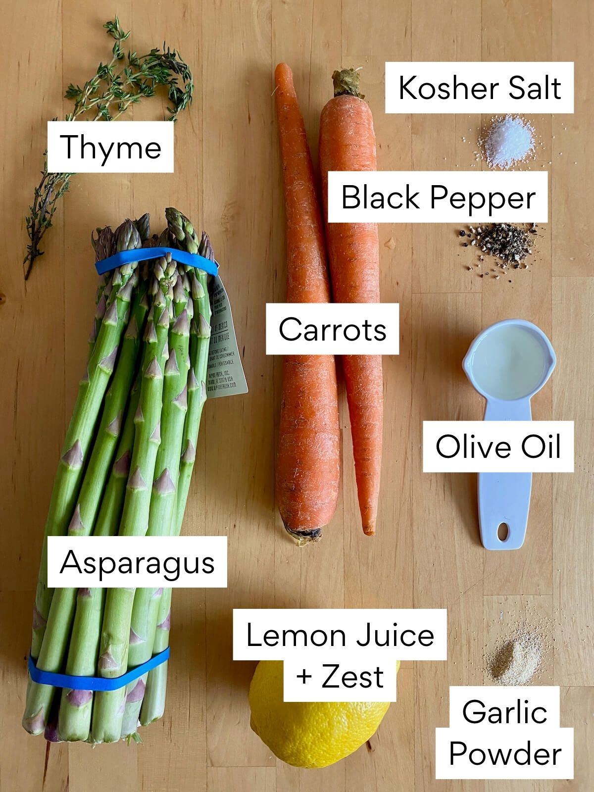 The ingredients to make roasted carrots and asparagus. Each ingredient is labeled with text. They include thyme, kosher salt, black pepper, carrots, asparagus, olive oil, lemon juice and zest, and garlic powder.