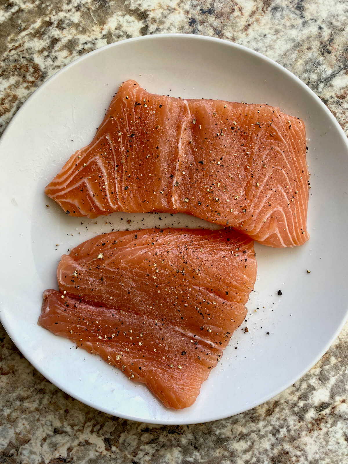 Two raw salmon fillets seasoned with salt and pepper on a white plate.