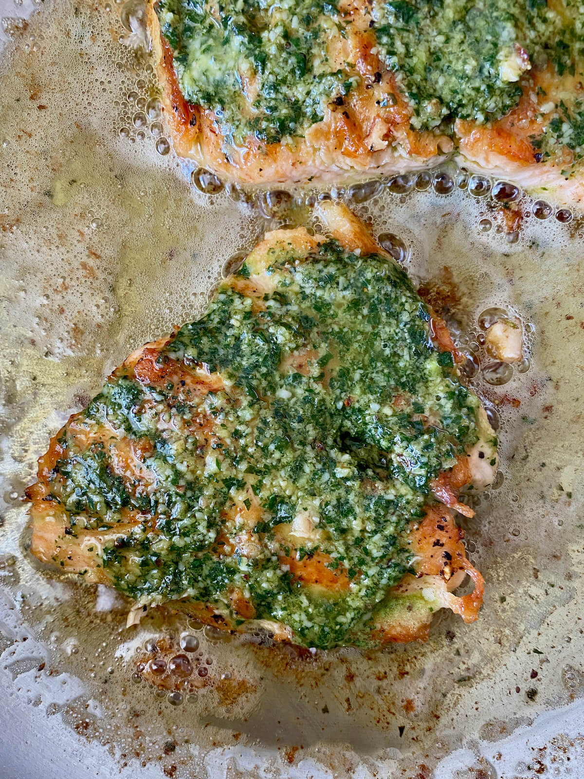 Salmon coated with pesto butter cooking in a stainless steel skillet.