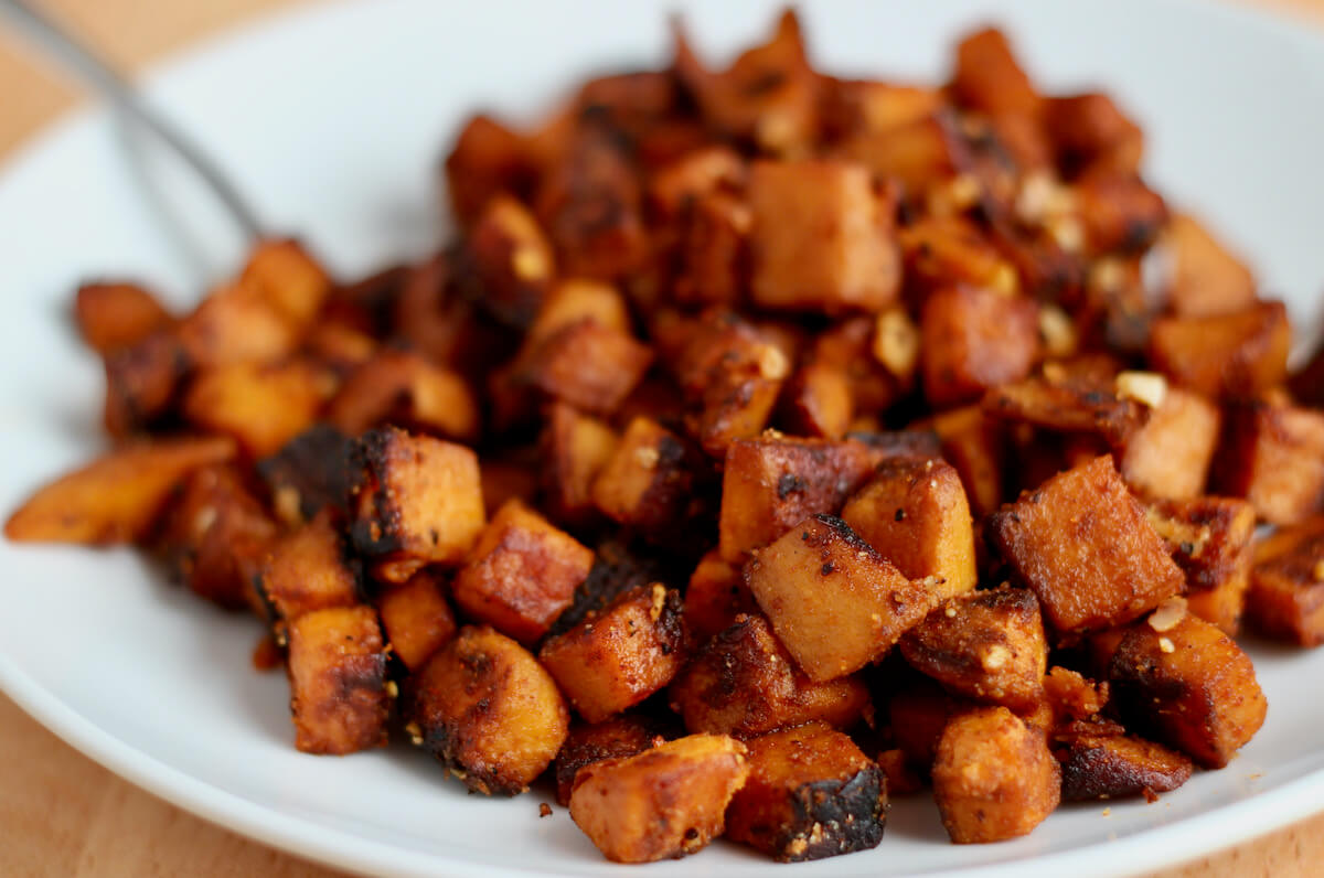 Sautéed sweet potatoes on a small white plate. There is a fork sticking out of the potatoes out of focus in the background.