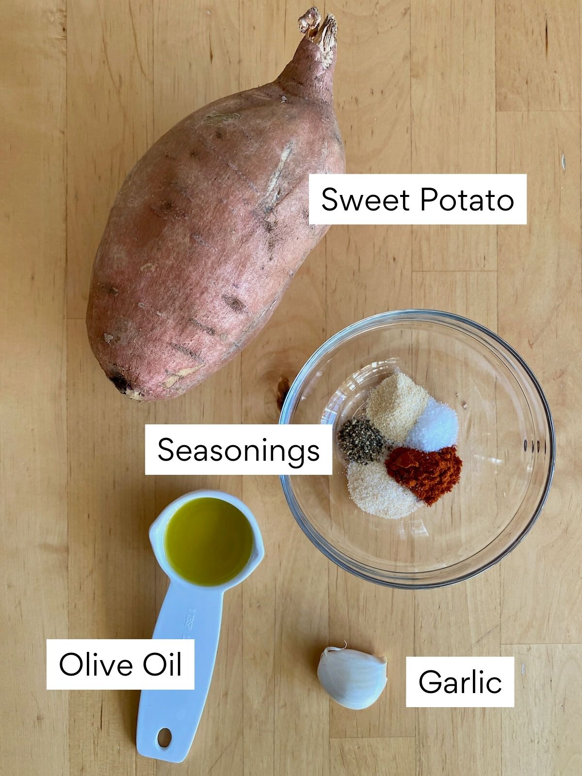 The ingredients to make sautéed sweet potatoes on a butcher block countertop. Each ingredient is labeled with text. They include sweet potato, seasonings, olive oil, and garlic.