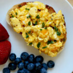 Scrambled eggs on toast on a small white plate with fresh fruit.