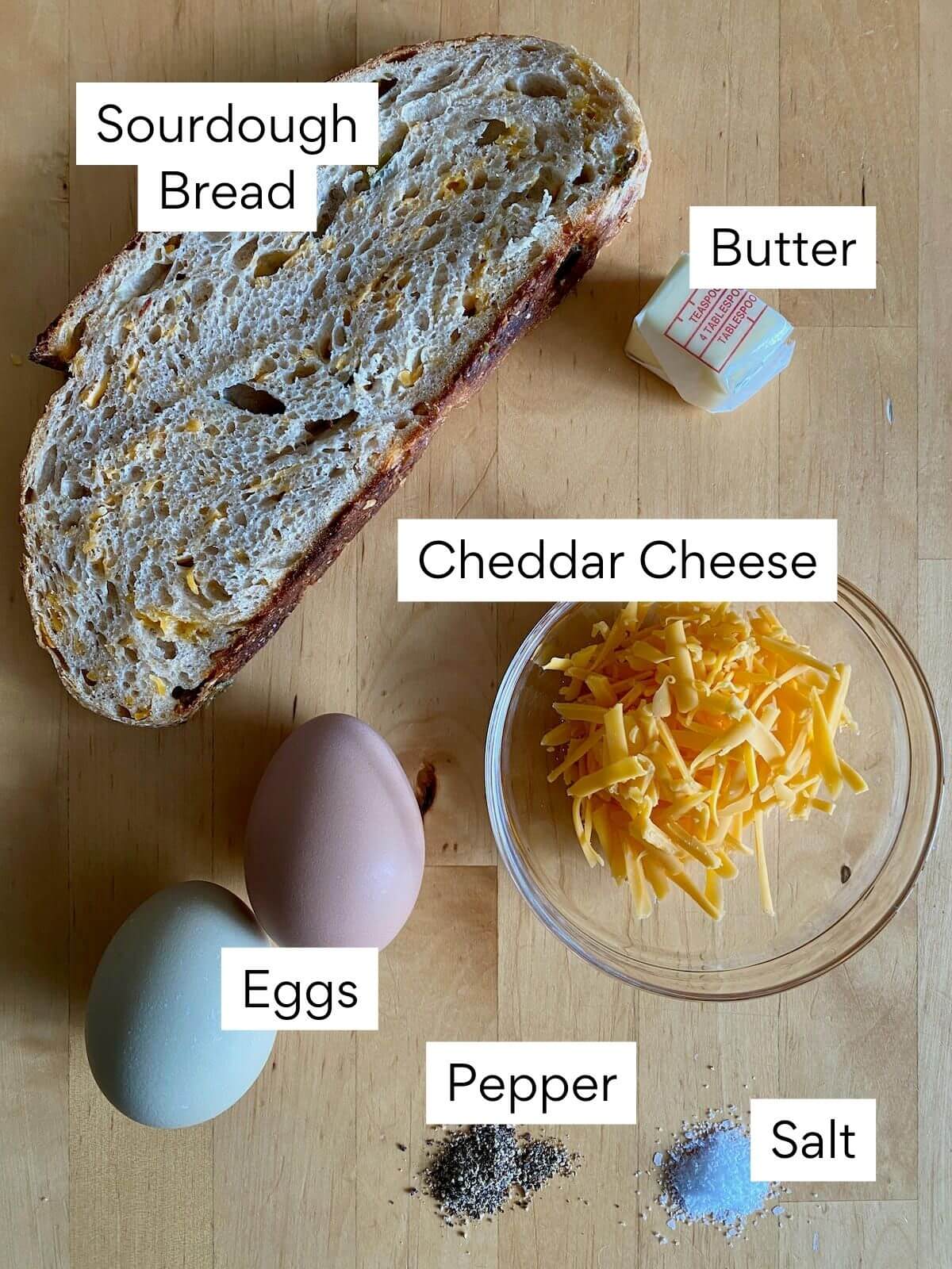 The ingredients to make scrambled eggs on toast. Each ingredient is labeled with text. They include sourdough bread, butter, cheddar cheese, eggs, pepper, and salt.