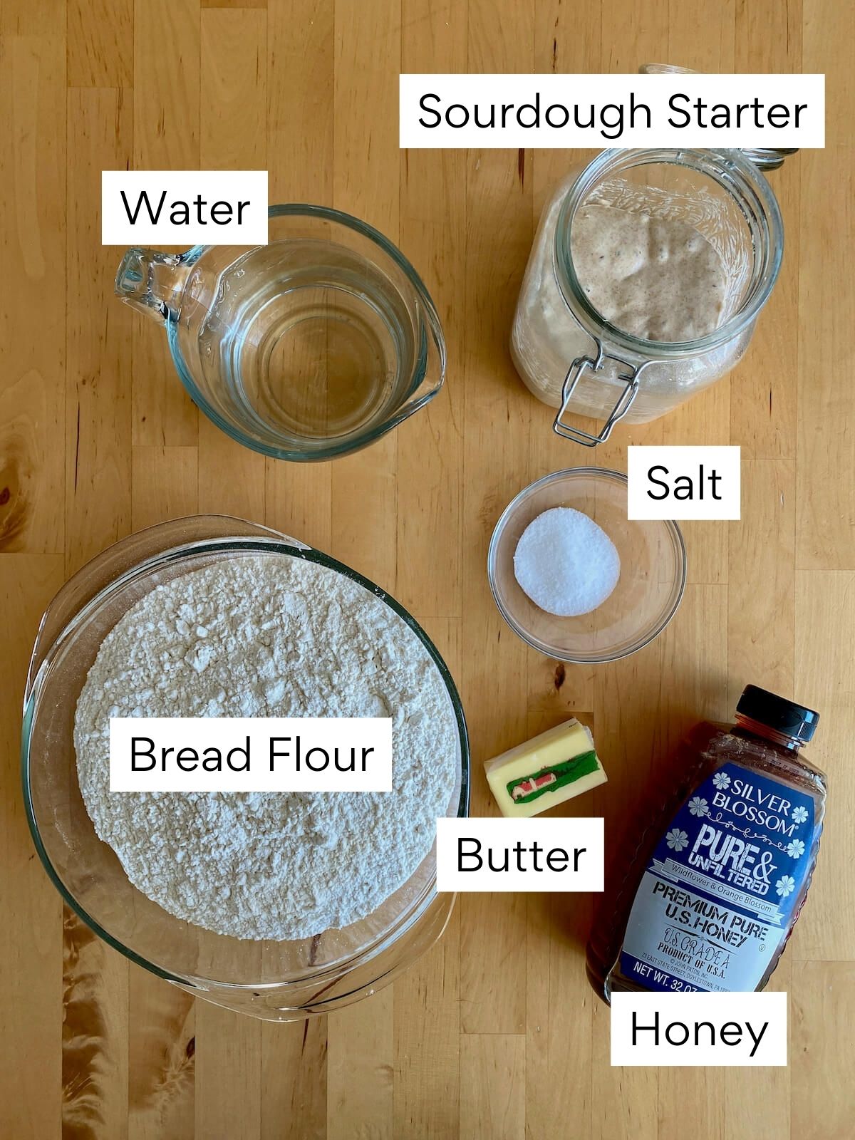 The ingredients to make sourdough hoagie rolls on a butcher block countertop. Each ingredient is labeled with text. They include sourdough starter, water, bread flour, salt, butter, and honey.