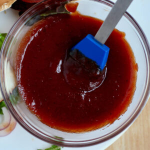 A bowl of apricot bbq sauce with a blue basting brush in it. The bowl is next to some burgers on a plate.