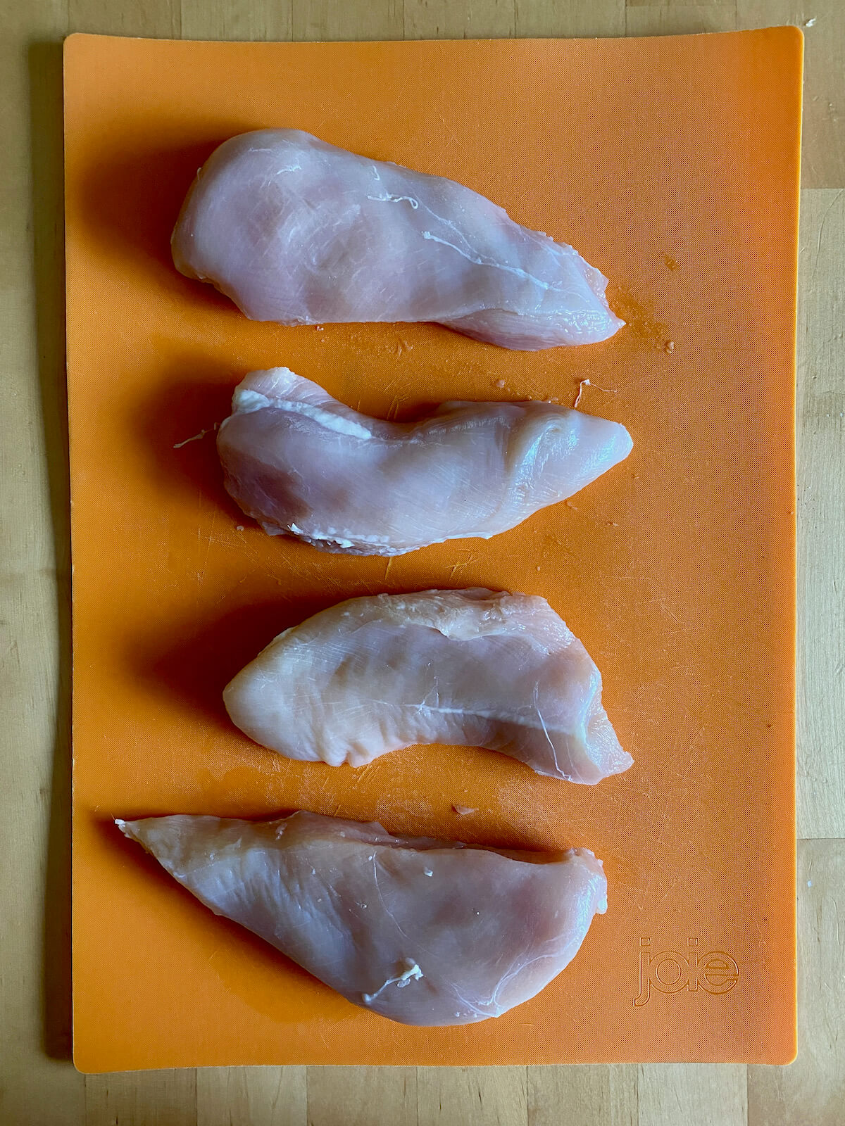 Four trimmed and cut chicken breasts on an orange cutting board.