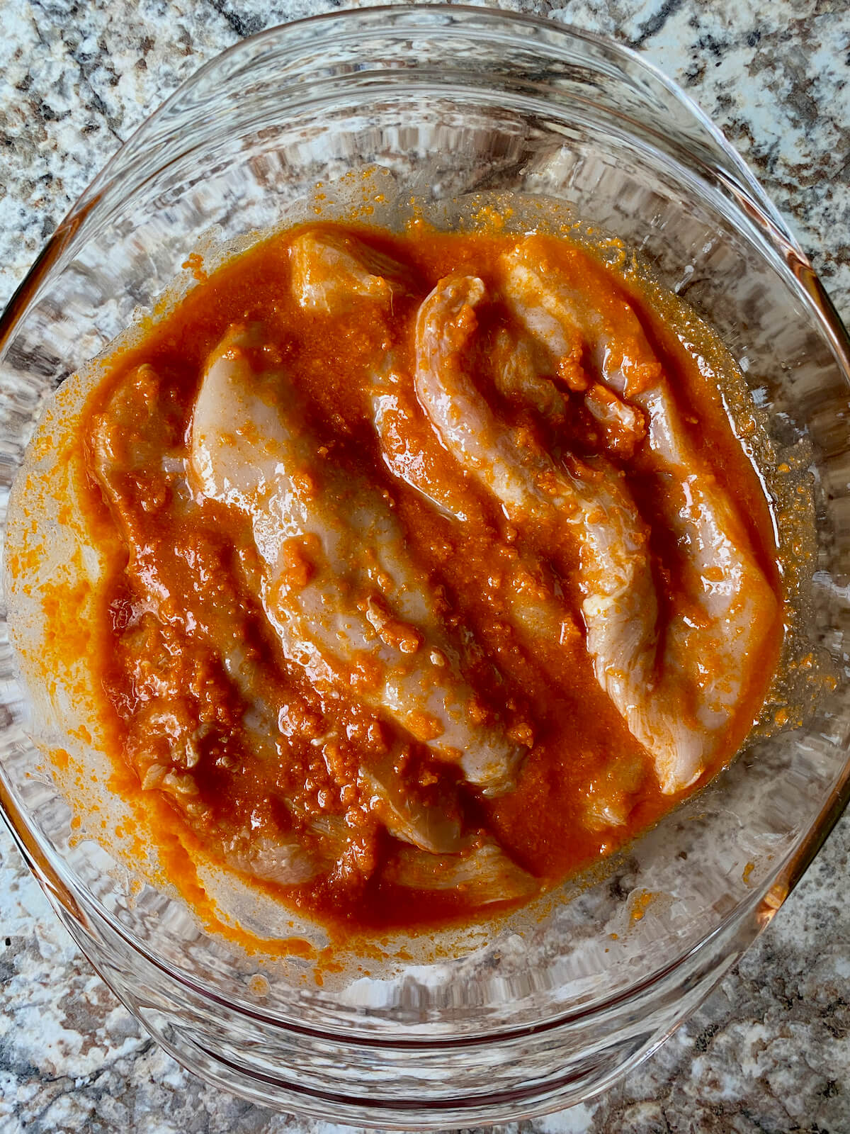 Chicken breasts marinating in homemade buffalo sauce in a glass bowl.