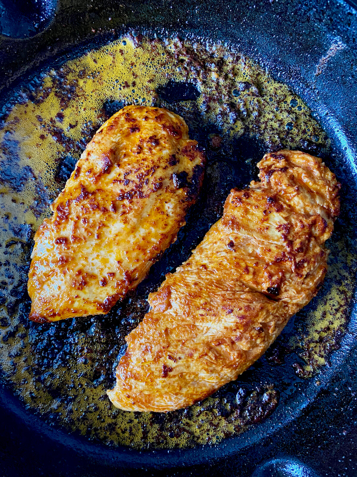 Two buffalo chicken breasts searing in a cast iron skillet.