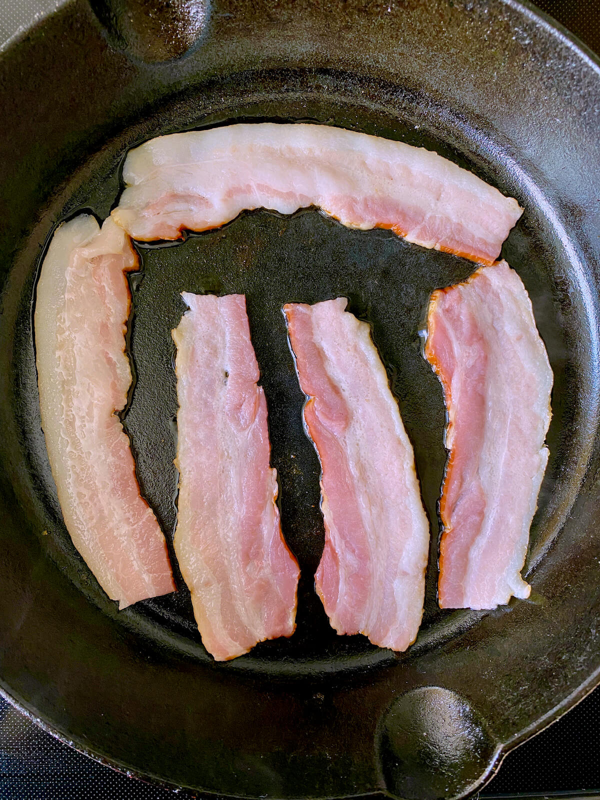 Bacon starting to cook in a cast iron skillet on the stovetop.