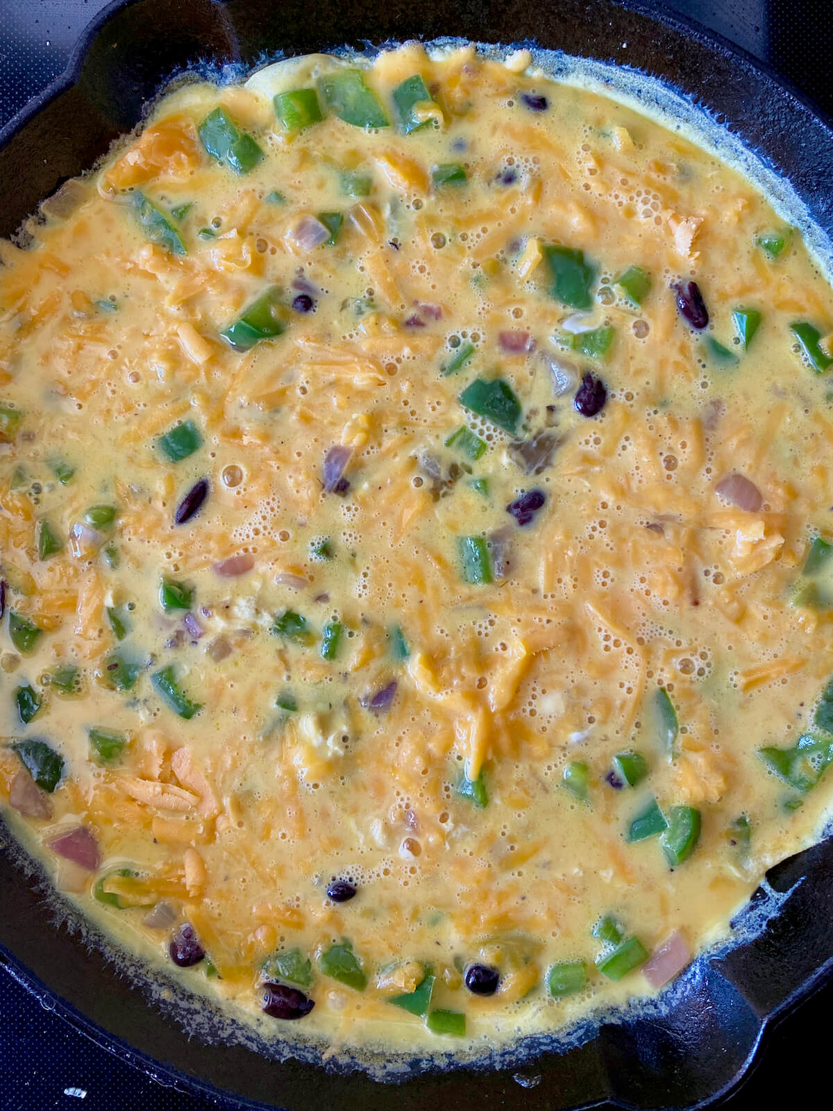 Raw eggs, sautéed vegetables, and cheddar cheese in a cast iron skillet on the stovetop. The edges of the egg mixture are just set.