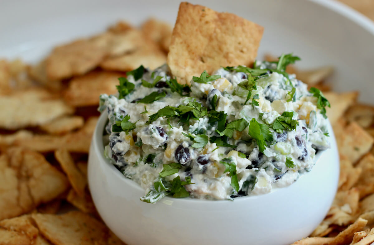 Black bean corn feta dip in a small white serving bowl with a pita chip sticking out of it. The small serving bowl is inside of a larger serving bowl filled with pita chips.