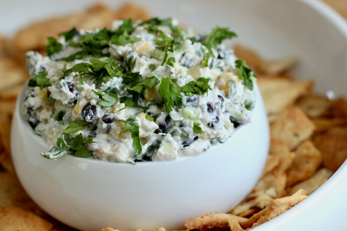 Feta black bean corn dip in a small white serving bowl inside of a larger serving bowl filled with pita chips.