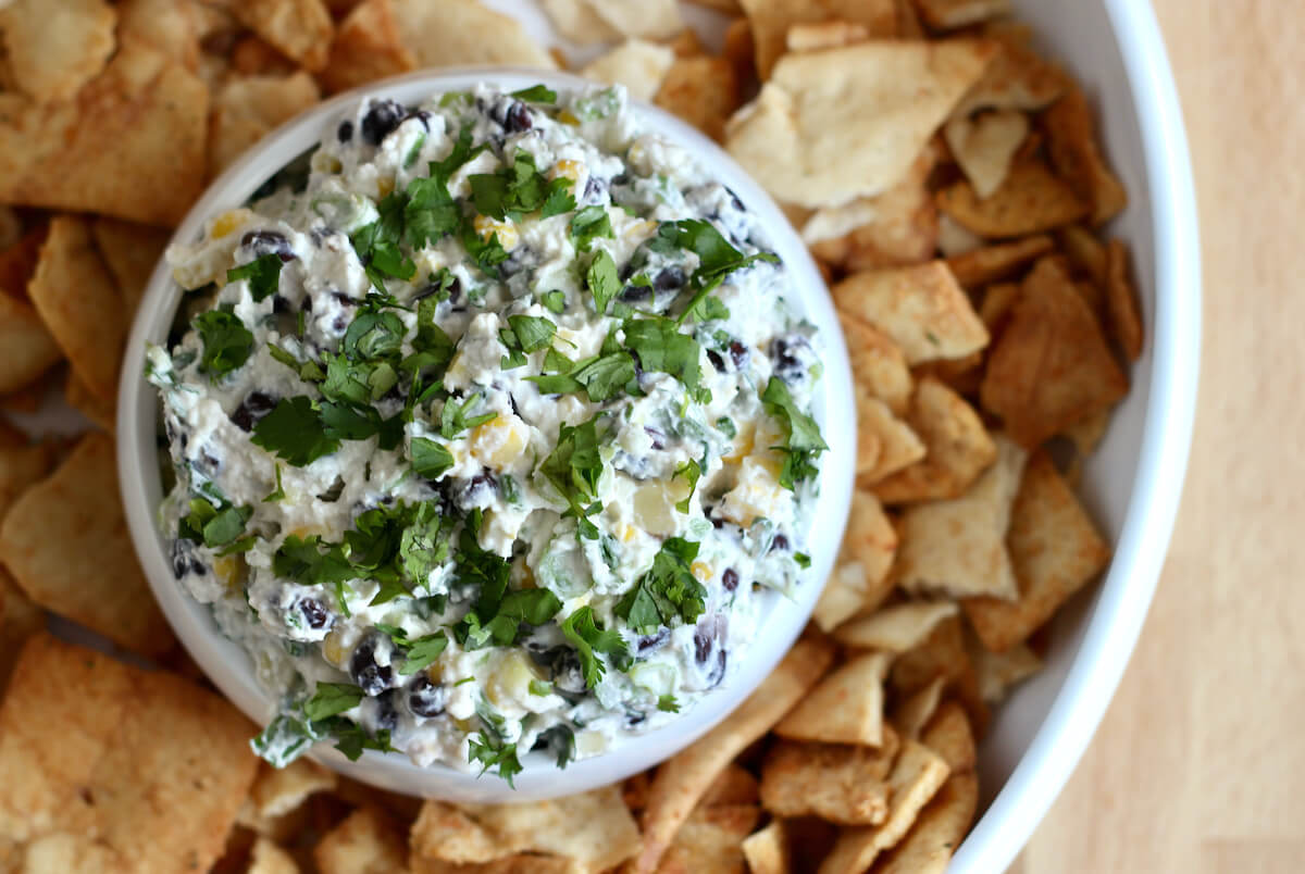 A bowl of corn black bean feta dip inside of a large serving bowl filled with pita chips.