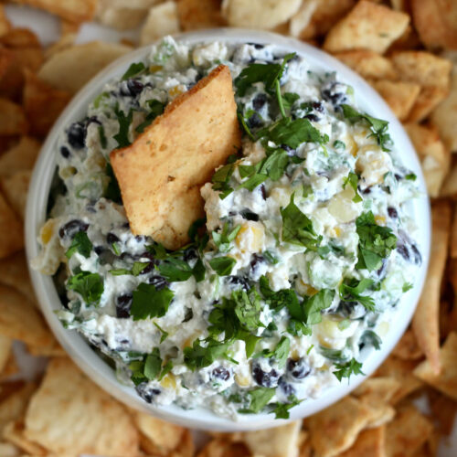A bowl of black bean corn feta dip surrounded by pita chips. There is a pita chip sticking out of the dip.