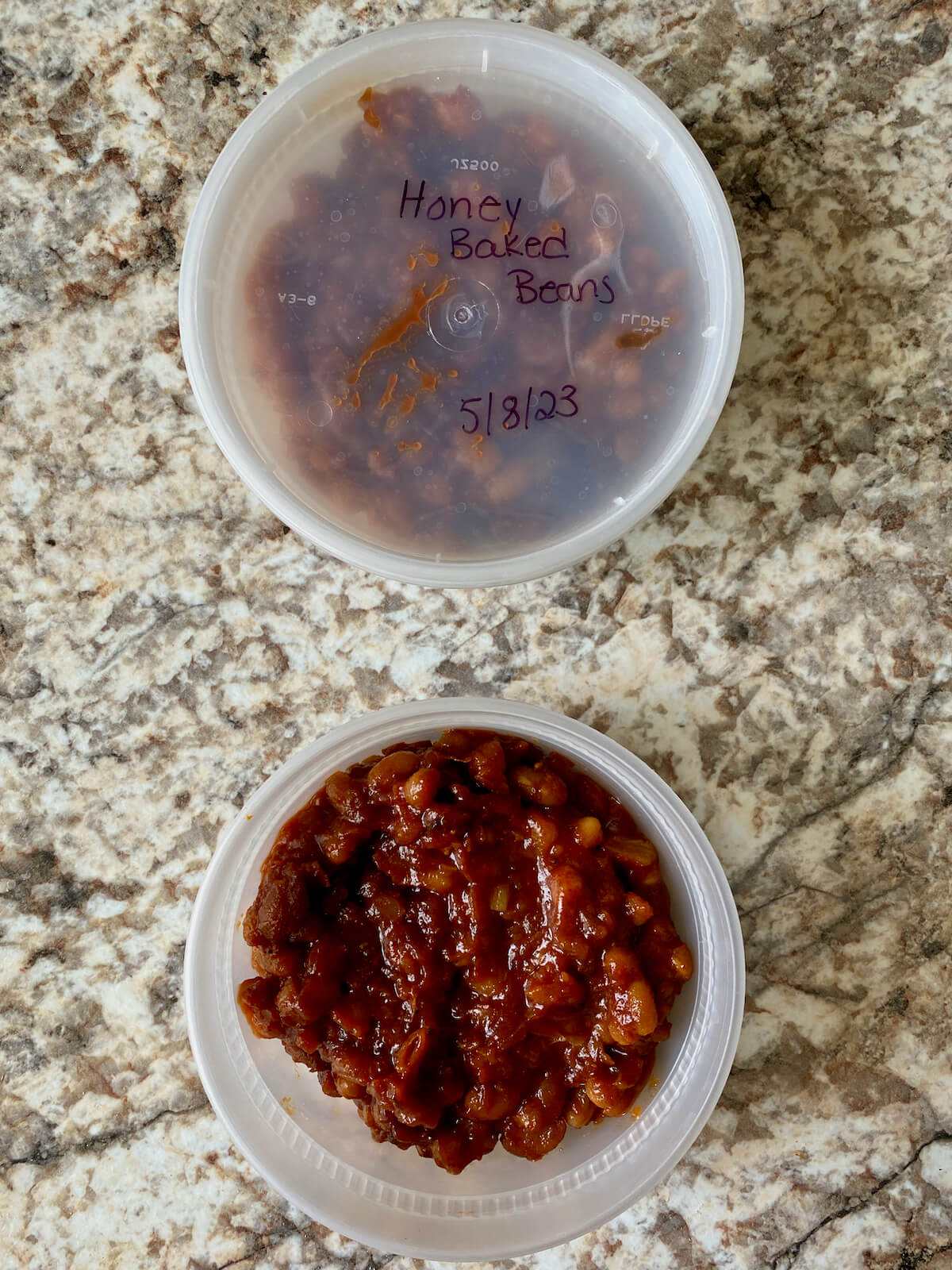 Two round plastic storage containers filled with homemade baked beans. One container has a lid on it that's labeled "Honey Baked Beans, 5/8/23."