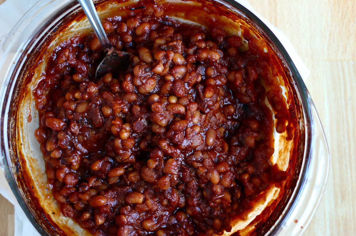 A round glass casserole dish filled with honey baked beans. There is a large serving spoon sticking out of the beans to the left.