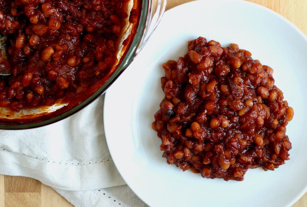 A small white plate full of honey baked beans next to a casserole dish filled with the rest of the baked beans.