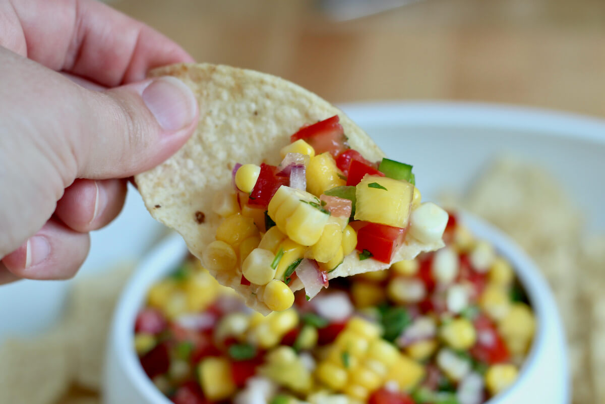 A hand holding up a tortilla chip with some mango corn salsa on it.
