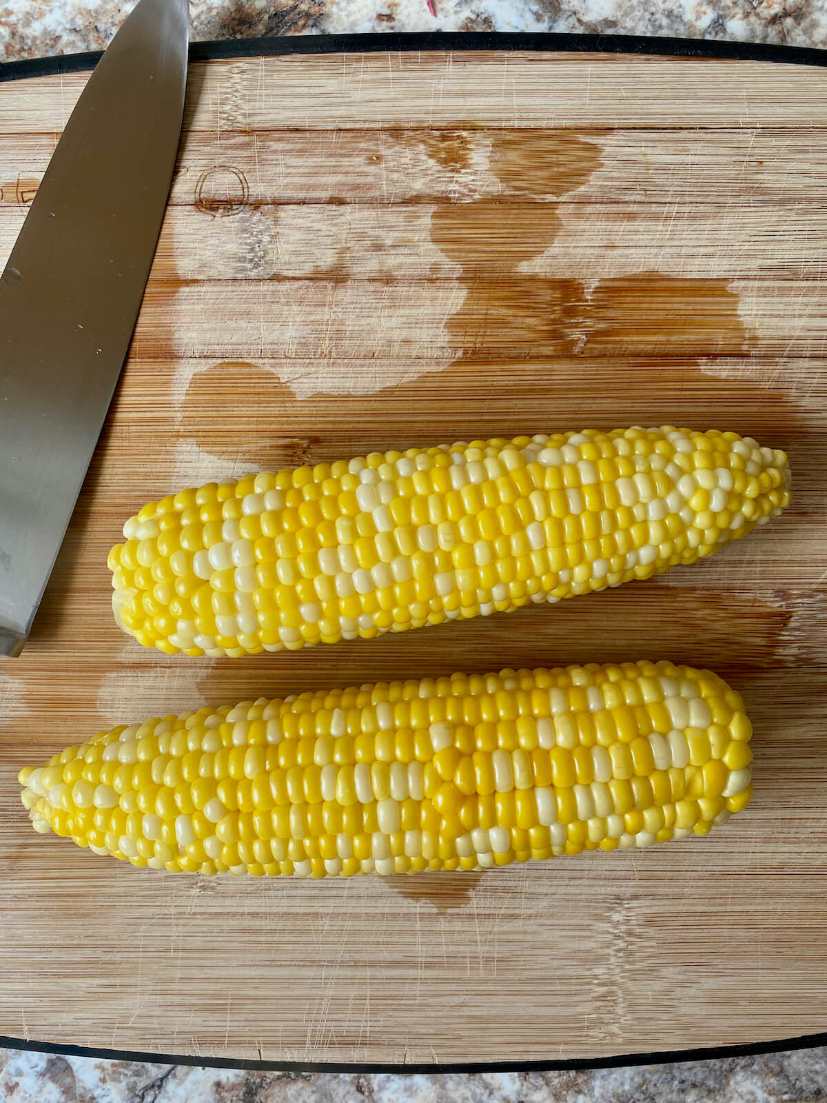 Two cooked ears of fresh corn on the cob on a bamboo cutting board next to a knife.