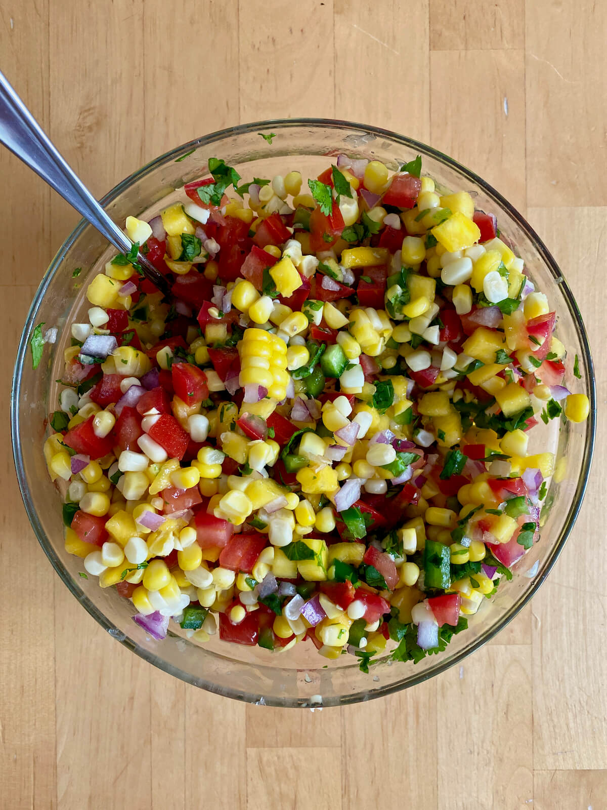 The prepared ingredients to make mango corn salsa mixed together in a clear glass bowl with a silver spoon sticking out of the bowl to the left.