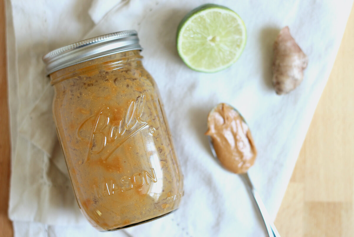 A mason jar of peanut butter laying on a white cloth napkin next to half a lime, a knob of fresh ginger, and a spoonful of peanut butter.