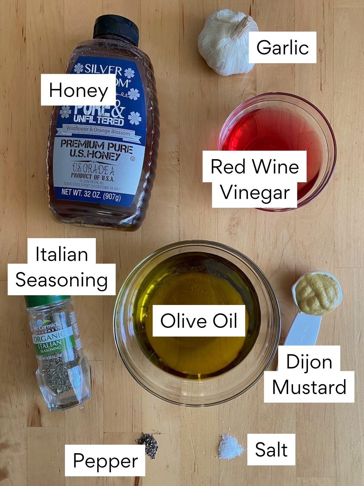 The ingredients to make red wine vinegar salad dressing. Each ingredient is labeled with text. They include honey, garlic, red wine vinegar, Italian seasoning, olive oil, dijon mustard, pepper, and salt.