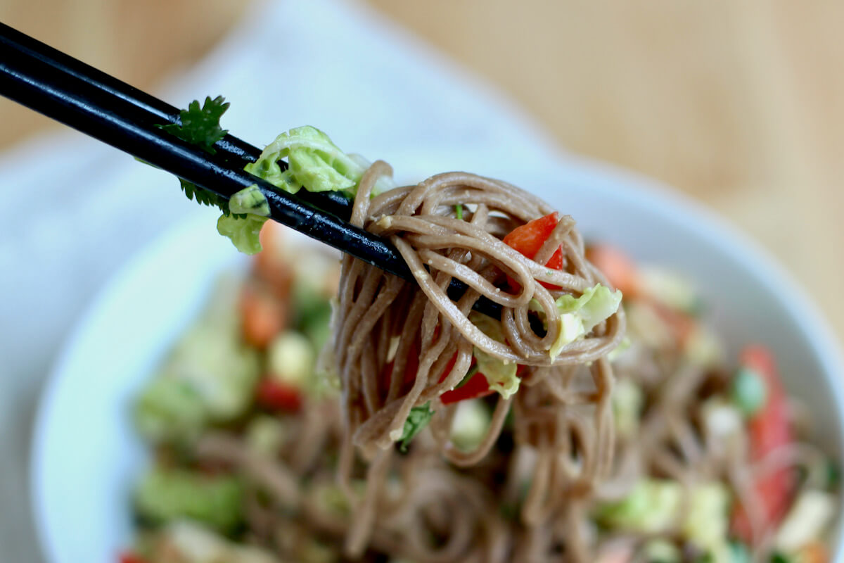 Black chopsticks holding up a bite of cold soba noodle salad above a serving bowl.