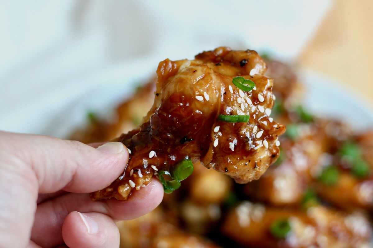 A hand holding a soy garlic chicken wing over a plate of chicken wings.