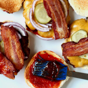 Several open-faced bbq bacon burgers on a white plate. There is a basting brush brushing bbq sauce on one of the buns.