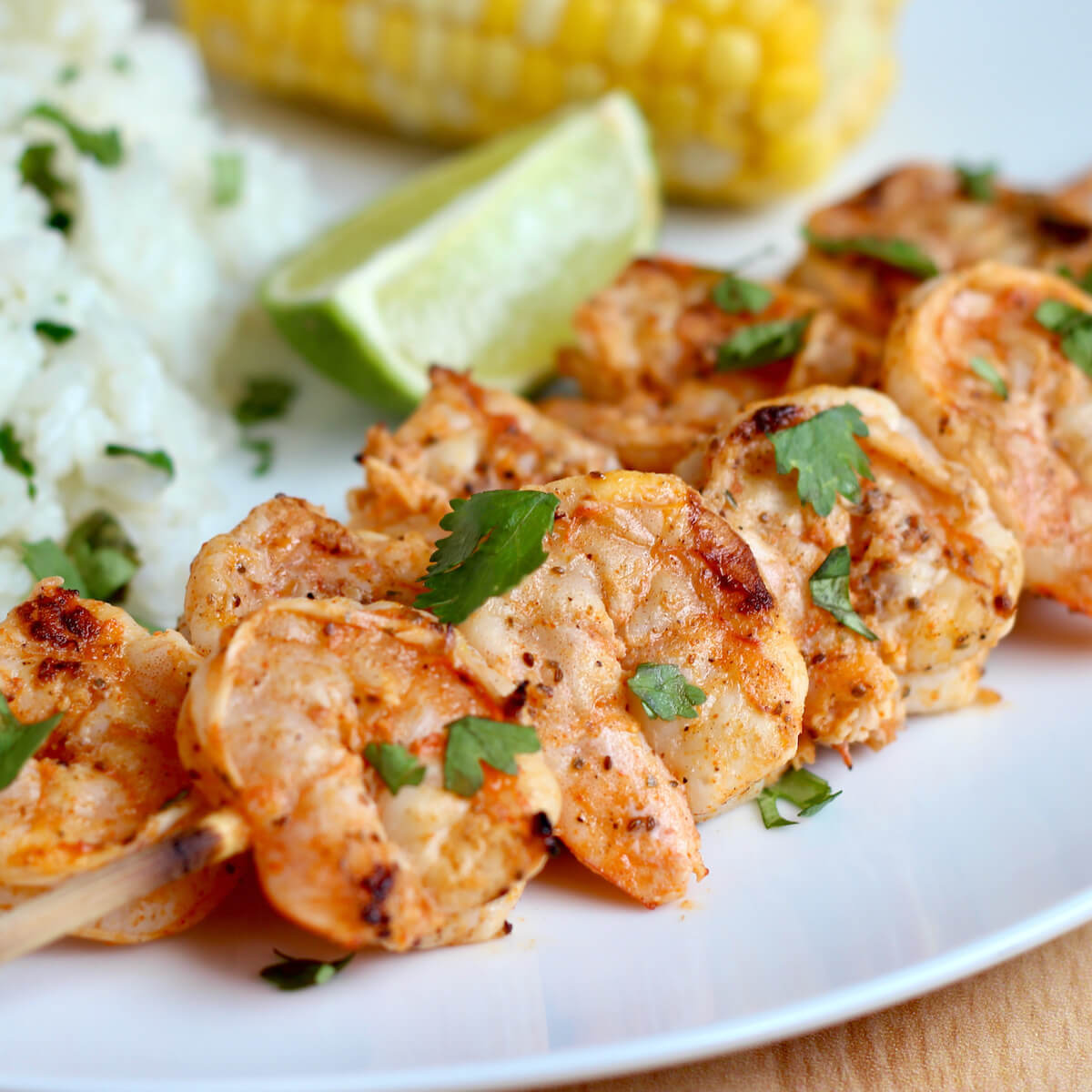 Two skewers of Old Bay grilled shrimp on a white plate with rice and corn on the cob.