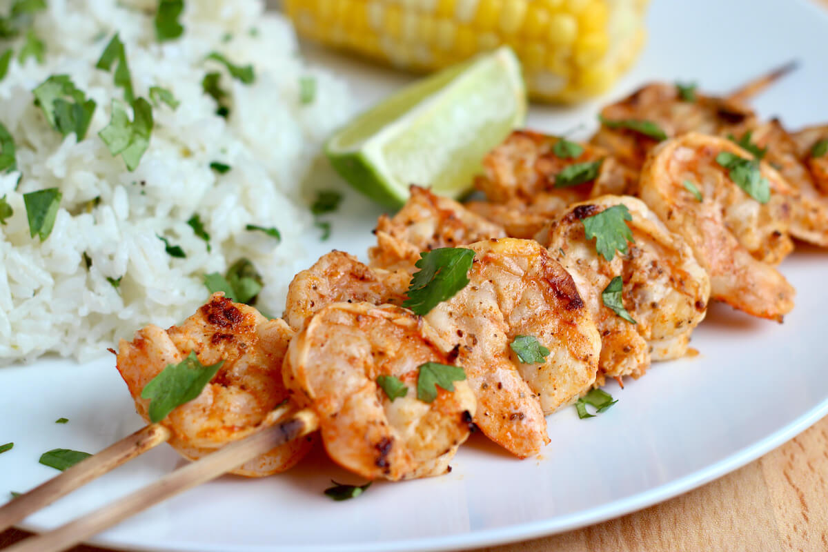 Two skewers of Old Bay grilled shrimp on a white plate with rice and corn on the cob.