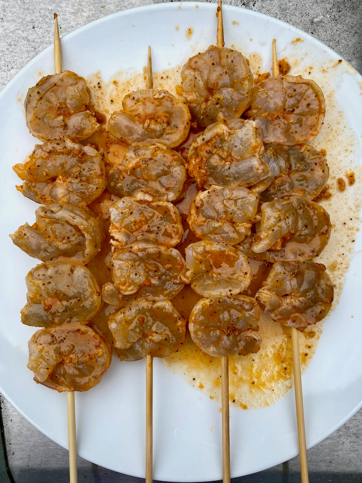 Four skewers of marinated raw shrimp on a white plate.