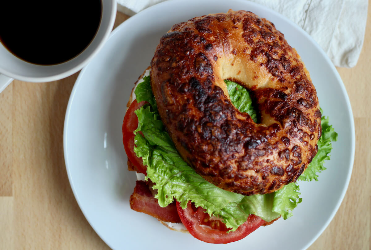 A BLT sandwich on a bagel on a small white plate next to a cup of black coffee.