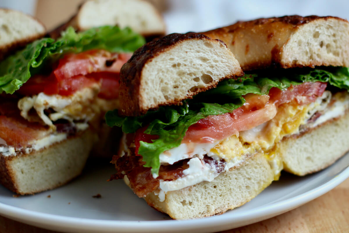 A BLT sandwich on a bagel with egg cut in half on a small white plate.