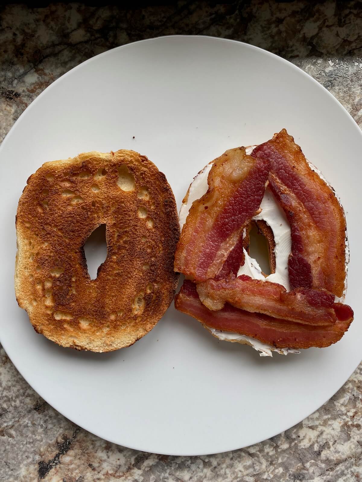 Bacon slices and cream cheese on one half of a bagel on a white plate.