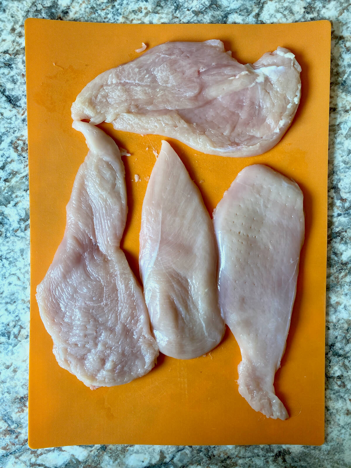 Four chicken breasts pounded out on an orange cutting board.