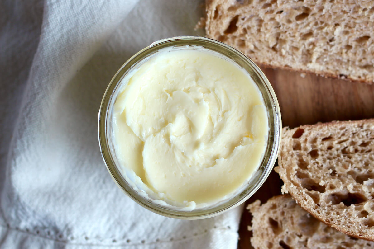 A small glass jar of butter next to slices of sourdough bread. There is a white cloth napkin laying beneath the jar of butter.