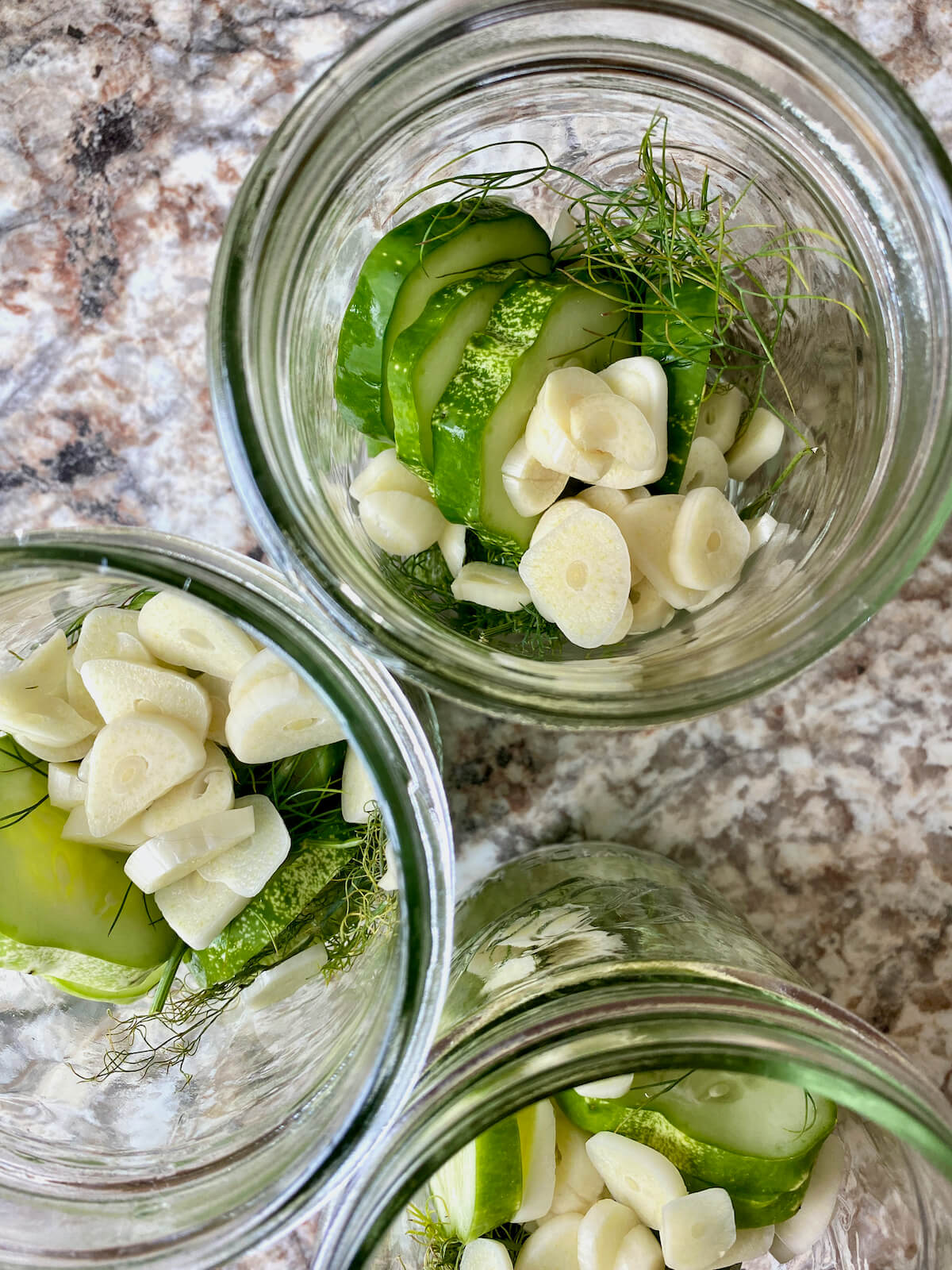 Sliced cucumbers, sliced garlic, and fresh dill in glass mason jars.