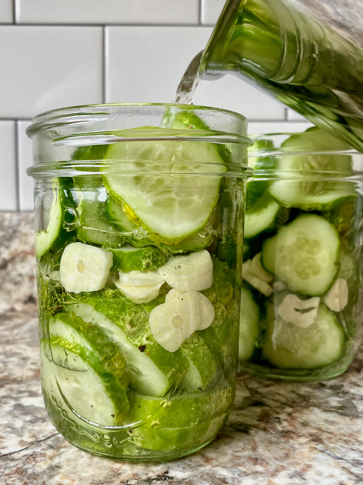 Brining liquid being poured from a stainless steel pot into mason jars filled with cucumber slices.